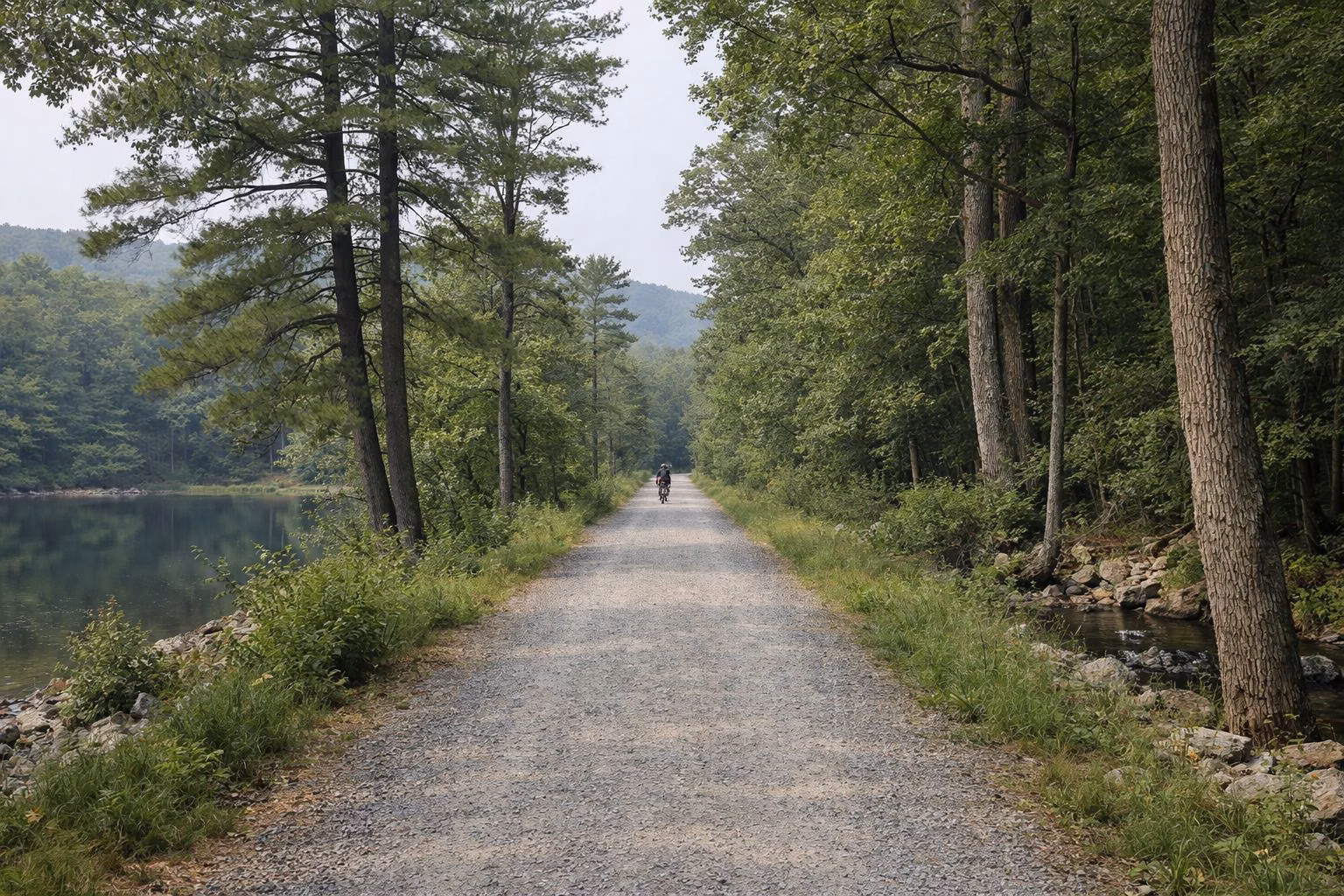 A wide flat crushed-stone trail running straight through green woods, a cyclist far in the distance