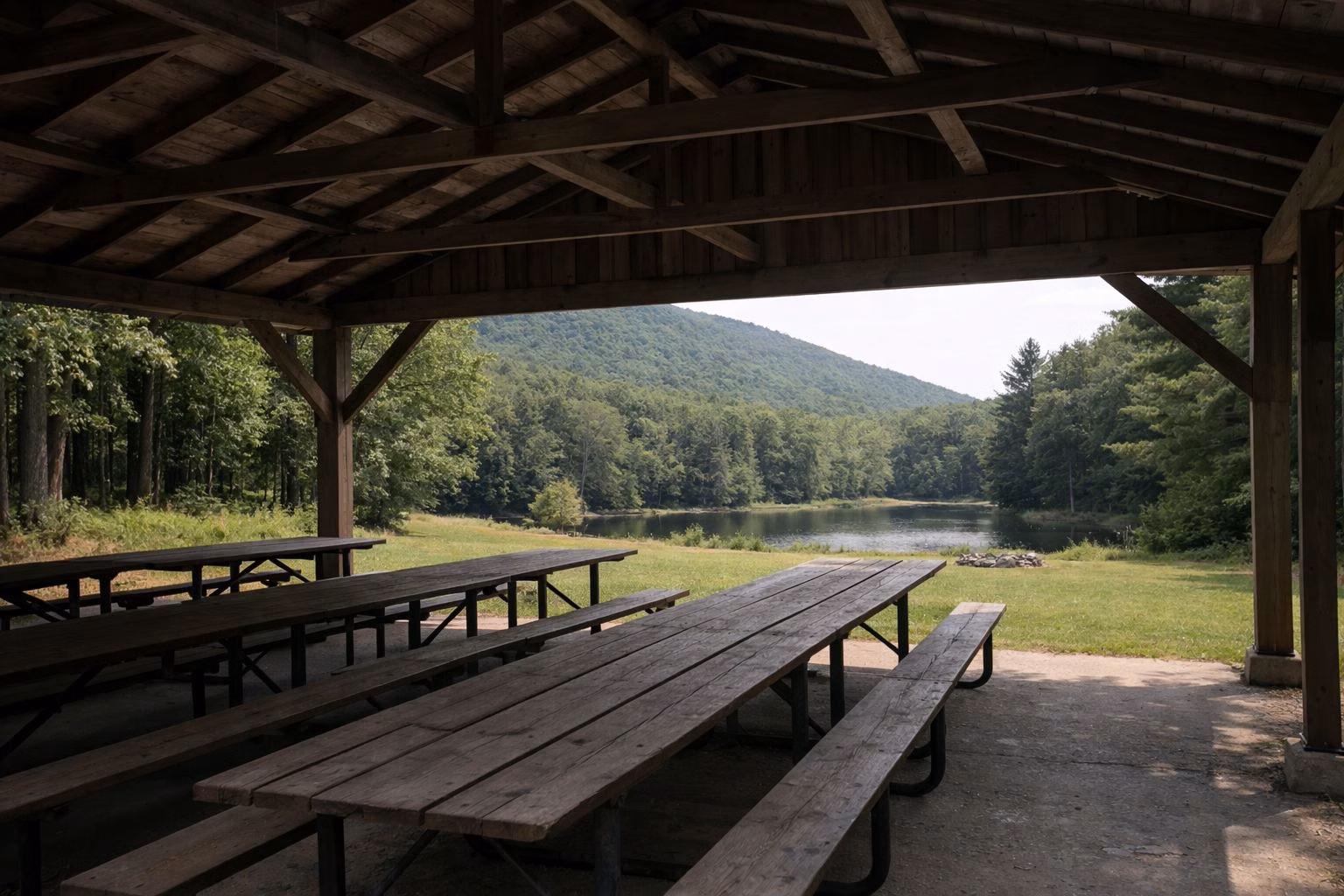 An open-sided wooden picnic pavilion with long picnic tables at the edge of oak woods and a mown lawn