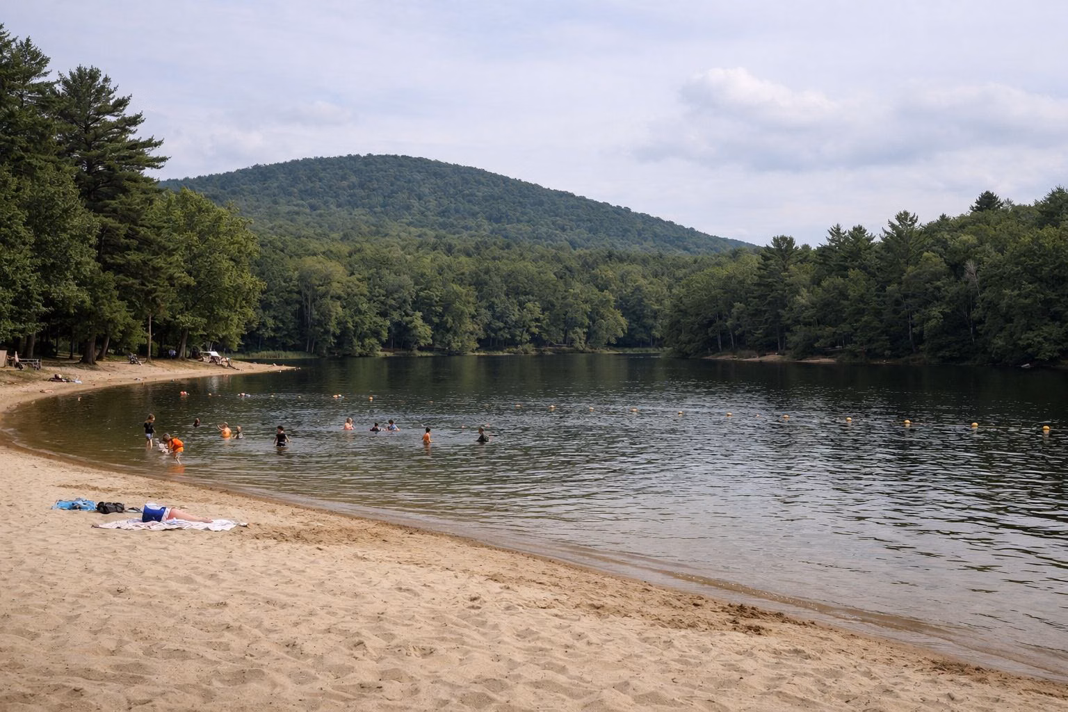 A small sandy lake beach with shallow water and a few distant swimmers, forest around the shore